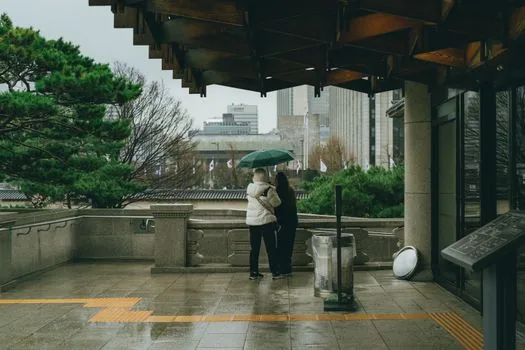 smartphone showing disaster safety app settings beside umbrella and raincoat near apartment entrance, Korean rainy season mood, realistic editorial photo, no readable private data